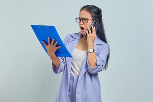 Portrait of surprised young Asian woman talking on mobile and looking at task list in documents folder isolated on purple background photo
