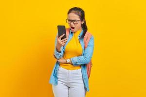 retrato de una joven estudiante asiática enojada vestida de forma informal con mochila usando un smartphone mientras gritaba en voz alta aislada en un fondo amarillo. educación en concepto de universidad universitaria