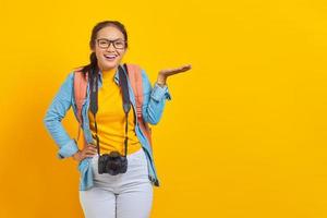 Portrait of cheerful young traveler Asian woman with backpack and camera in denim clothes while inviting to come on yellow background. Passenger traveling on weekends. Air flight journey concept photo