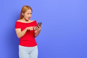 Portrait of surprised young Asian woman in red dress typing text message or scrolling feed on social network using smartphone isolated over purple background photo