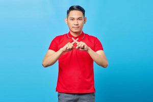 Portrait of serious handsome young man in red shirt crossing fingers and looking at camera with showing rejection attitude on blue background photo