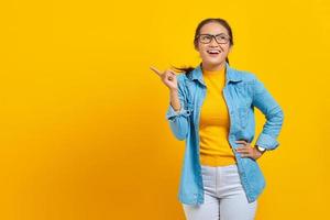 retrato de una alegre joven estudiante asiática vestida de denim señalando con el dedo en el espacio de copia, mostrando productos publicitarios aislados en un fondo amarillo. educación en concepto de universidad universitaria foto