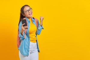 retrato de una alegre joven estudiante asiática vestida de forma informal con mochila usando un teléfono móvil y celebrando el éxito, recibiendo buenas noticias aisladas en un fondo amarillo