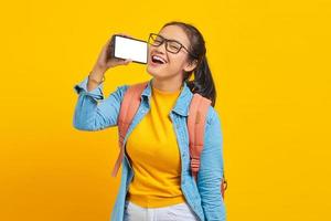 Portrait of smiling young Asian woman student in casual clothes with backpack enjoying singing using smartphone isolated on yellow background. Education in college university concept photo