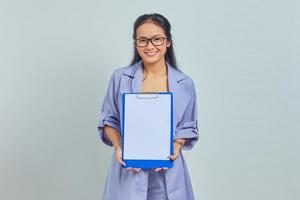 Portrait of cheerful young Asian business woman showing blank clipboard while looking at camera isolated on purple background photo