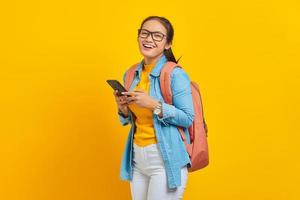 Portrait of cheerful young Asian woman student in casual clothes with backpack using mobile phone and looking at camera isolated on yellow background. Education in college university concept photo