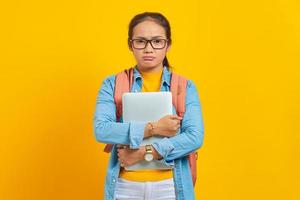 retrato de una joven estudiante asiática disgustada con ropa informal con mochila sosteniendo una laptop y mirando una cámara aislada en un fondo amarillo. educación en concepto de colegio universitario