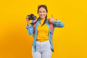 retrato de una joven asiática sonriente con mochila sosteniendo una cámara profesional y mostrando un gesto con el pulgar hacia arriba aislado en un fondo amarillo foto