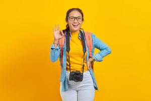Portrait of cheerful young traveler Asian woman with backpack and camera in denim clothes showing okay gesture on yellow background. Passenger traveling on weekends. Air flight journey concept photo