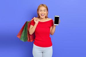 Cheerful beautiful Asian woman in red dress holding shopping bags, showing blank screen phone isolated over purple background photo