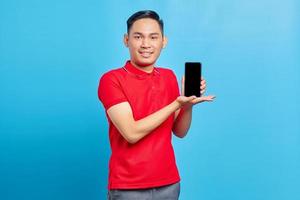 Portrait of smiling young asian man in red shirt showing blank screen mobile phone and looking at camera isolated on blue background photo