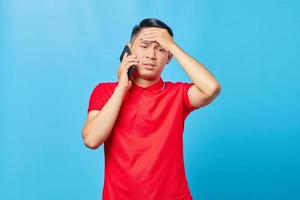 Portrait of dizzy asian man talking on smartphone and holding head with palm isolated on blue background photo