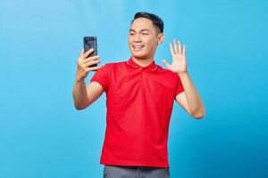 Portrait of cheerful Asian young man making video call and waving at smartphone camera isolated on blue background photo