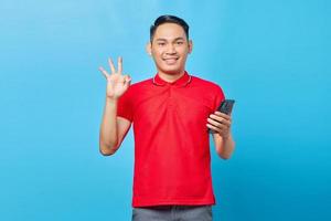 retrato de un joven asiático sonriente sosteniendo un teléfono móvil y haciendo un gesto de aprobación aislado en el fondo azul foto
