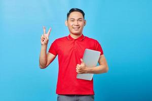 Portrait of smiling Asian young man in red shirt holding laptop and showing peace sign with finger while looking at camera isolated on blue background photo