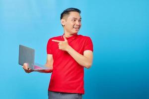 Portrait of cheerful Asian young man in red shirt pointing at laptop with finger isolated on blue background photo