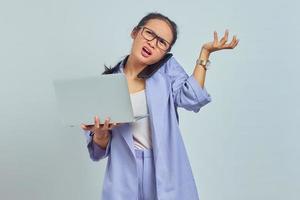 Portrait of angry young Asian woman talking on mobile phone and holding laptop, pointing to copy space with palms isolated on white background photo