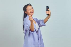 retrato de una alegre joven asiática usando un teléfono móvil tomando selfie y mostrando el signo del corazón con dos dedos cruzados aislado en fondo blanco foto