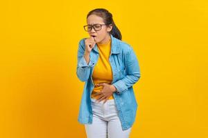 Beautiful young Asian woman student in denim clothes suffering with cough and feeling bad isolated on yellow background. Education in college university concept photo