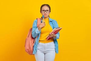 Portrait of serious young Asian woman student in casual clothes with backpack holding book and pen, thinking about question isolated on yellow background. Education in college university concept
