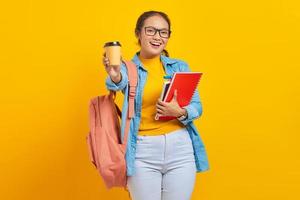 retrato de una alegre joven estudiante asiática vestida de forma informal con mochila sosteniendo un libro y mostrando café para llevar mirando una cámara aislada de fondo amarillo foto