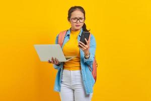 Confused young Asian woman student in casual clothes with backpack holding laptop and reading news message on smartphone isolated on yellow background. Education in college university concept