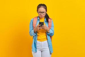 retrato de una joven estudiante asiática molesta vestida de forma informal con mochila usando un teléfono móvil, escribiendo mensajes sms aislados en un fondo amarillo. educación en concepto de universidad universitaria foto