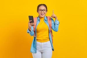 Portrait of  cheerful young Asian woman student in casual clothes with backpack holding mobile phone and pointing up space with fingers isolated on yellow background photo