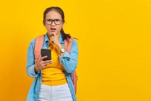 retrato de una joven estudiante asiática pensativa vestida de forma informal con mochila usando un teléfono móvil y sosteniendo la barbilla mientras piensa en algo aislado de fondo amarillo foto