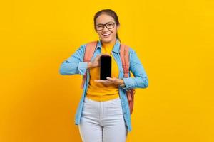 retrato de una alegre joven estudiante asiática vestida de forma informal con mochila que muestra un teléfono móvil con pantalla en blanco aislado en un fondo amarillo. educación en concepto de universidad universitaria foto