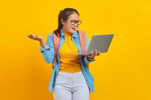 Portrait of smiling young Asian woman student in casual clothes with backpack showing copy space on palm and holding laptop isolated on yellow background. Education in university college concept