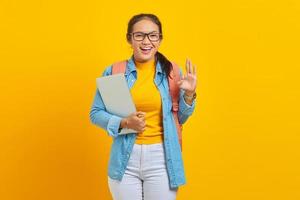 Portrait of cheerful young Asian woman student in casual clothes with backpack holding laptop and showing ok gesture with finger isolated on yellow background. Education in university college concept