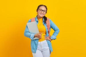 Portrait of cheerful young Asian woman student in casual clothes with backpack  holding a laptop and looking at camera isolated on yellow background. Education in university college concept photo