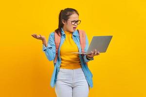 Portrait of annoyed young Asian woman student in casual clothes with backpack showing copy space on palm and holding laptop isolated on yellow background. Education in university college concept photo