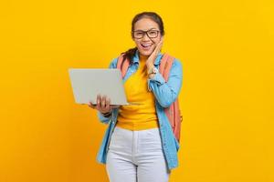 Portrait of excited young Asian woman student in casual clothes with backpack using laptop and  touching cheek with hands isolated on yellow background. Education in college university concept photo