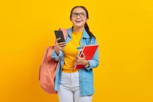 Portrait of smiling young Asian woman student in denim clothes, glasses with backpack holding mobile phone and books isolated on yellow background. Education in high school university college concept