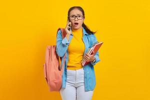 retrato de una joven estudiante asiática sorprendida vestida de denim con mochila hablando por teléfono móvil y sosteniendo un libro aislado de fondo amarillo foto