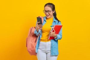 retrato de una alegre joven estudiante asiática vestida de denim con mochila usando un teléfono móvil escribiendo mensajes sms y sosteniendo libros aislados en un fondo amarillo foto