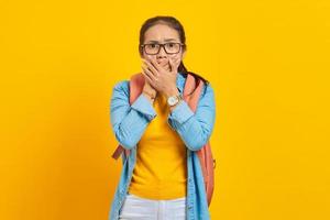 Shocked young Asian woman student in denim outfit with backpack covering mouth with hand isolated on yellow background. Education in university concept photo