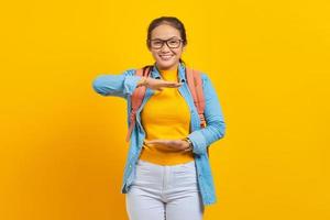 Cheerful young Asian woman student in denim outfit with backpack showing product with palm, showing copy space for certain discount isolated on yellow background. Education in university concept