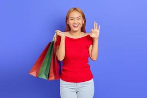 Cheerful beautiful Asian woman in red dress holding shopping bags, showing okay gesture isolated over purple background photo