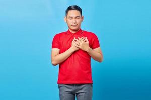 Portrait of handsome young Asian man in red shirt pressing hands to chest with closed eyes and grateful gesture on face. Health concept. photo