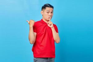 retrato de un apuesto joven asiático haciendo gestos de silencio y señalando el espacio de copia aislado sobre fondo azul