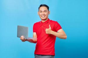 Portrait of cheerful Asian young man in red shirt using laptop and giving thumbs up, showing approval symbol isolated on blue background