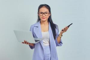 Portrait of young Asian woman standing using laptop and holding mobile phone with smiling expression looking at camera isolated on white background photo