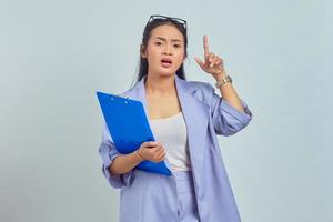 retrato de una joven asiática sorprendida sosteniendo una carpeta de documentos y señalando con el dedo hacia arriba aislada en un fondo morado foto