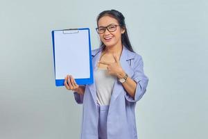 retrato de una joven y alegre mujer de negocios asiática que señala un portapapeles en blanco con el dedo aislado en un fondo morado foto