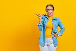 retrato de una alegre joven estudiante asiática vestida de denim señalando con el dedo en el espacio de copia, mostrando productos publicitarios aislados en un fondo amarillo. educación en concepto de universidad universitaria foto
