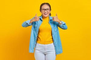 Portrait of smiling young Asian woman student in denim clothes  showing thumbs up gesture with hand isolated on yellow background. Education in college university concept photo