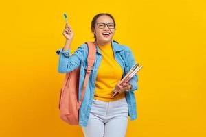 retrato de una alegre joven estudiante asiática vestida de forma informal con mochila sosteniendo un libro y señalando el espacio de copia con un bolígrafo aislado en un fondo amarillo. educación en concepto de universidad universitaria foto
